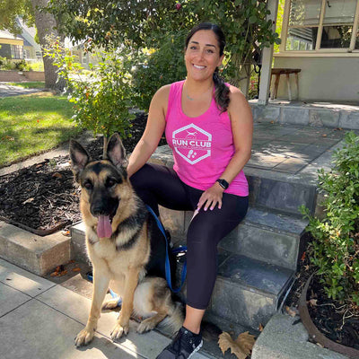 Woman wearing Canina Run Club women's tank top and running shoes while sitting with a German Shepherd