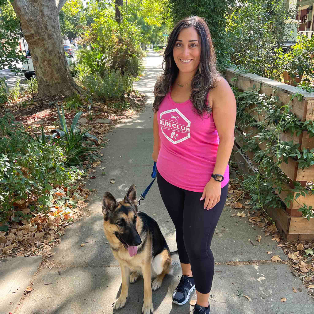 Woman wearing Canina Run Club women's tank top and running shoes while standing with a German Shepherd