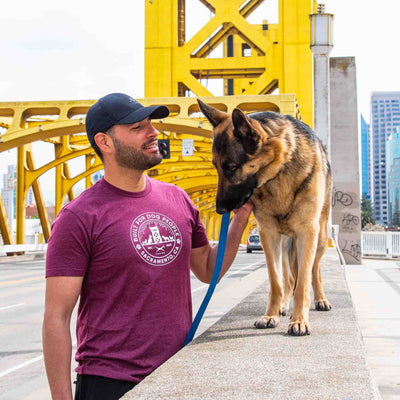 Man wearing Canina "Sactown" t-shirt standing with German Shepherd dog in front of bridge in Sacramento